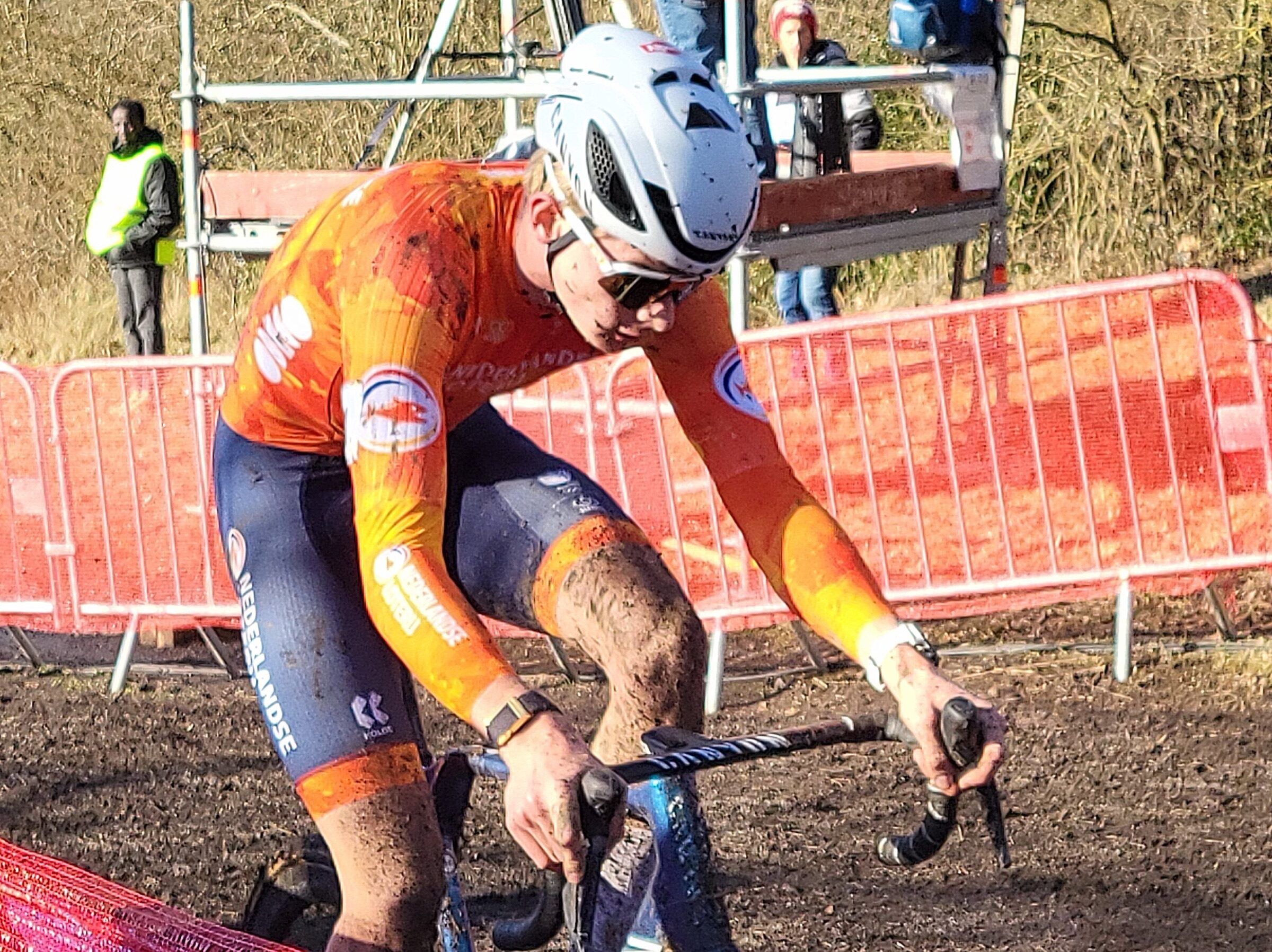 Tibor Del Grosso, maillot orange et casque blanc, les jambes couvertes de boue, roule le long des barrières rouges du parcours.
