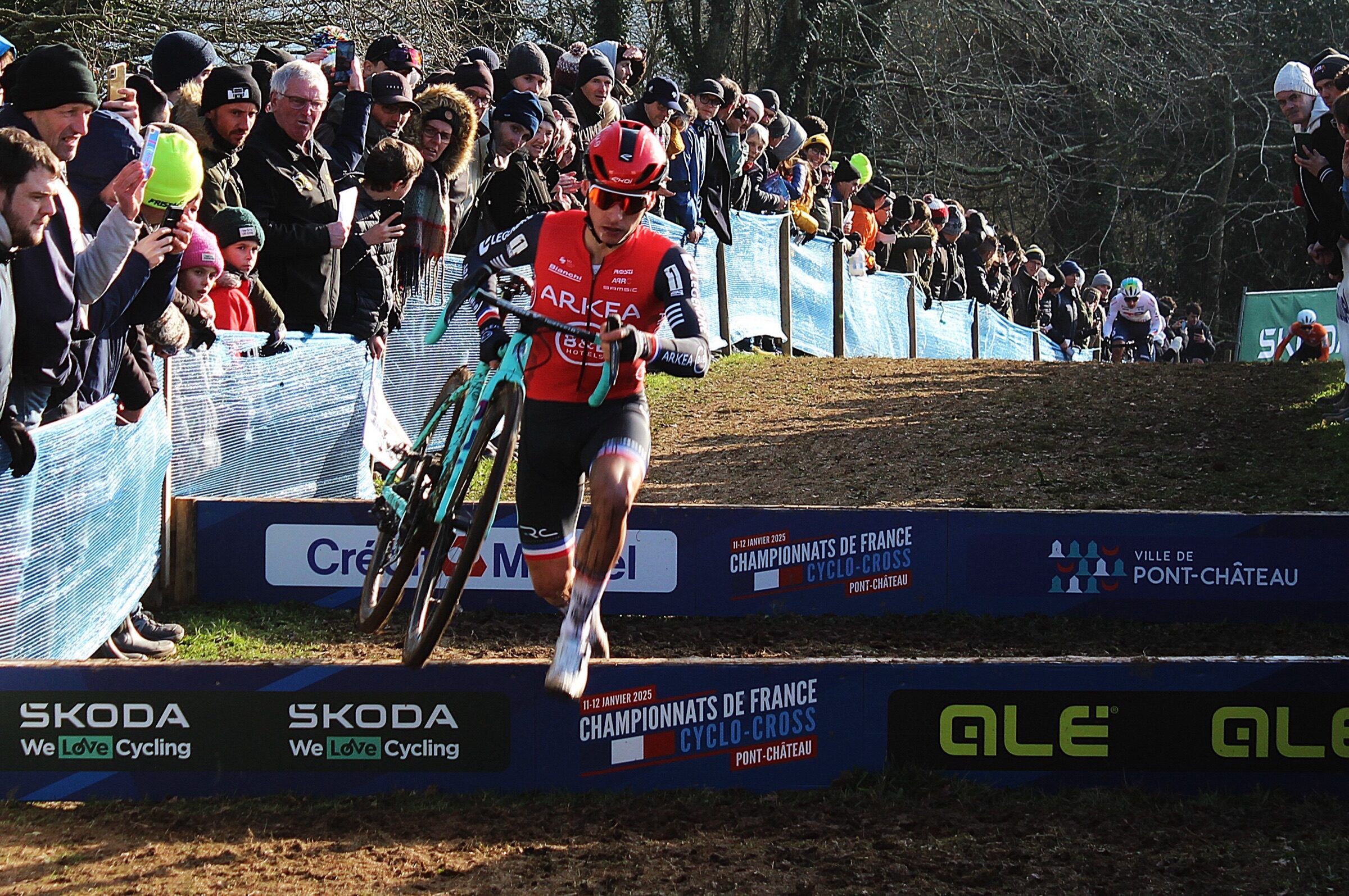 Un coureur en maillot rouge saute une planche en courant, vélo à la main, devant une foule massée le long des barrières bleues.