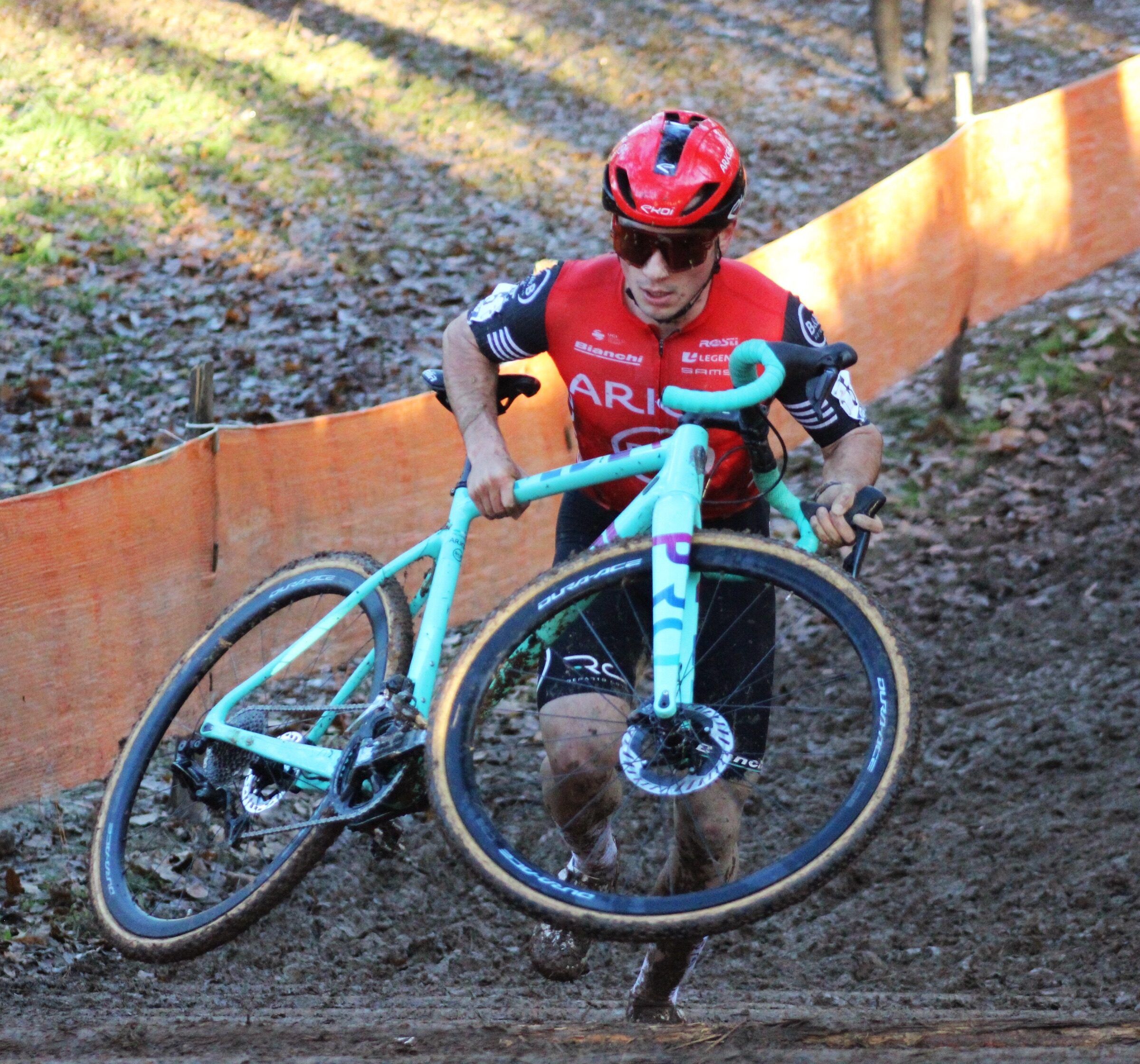 Rémi Lelandais, casque et maillot rouges, gravit une montée boueuse en portant son vélo vert d'eau devant lui, entre deux filets orange.